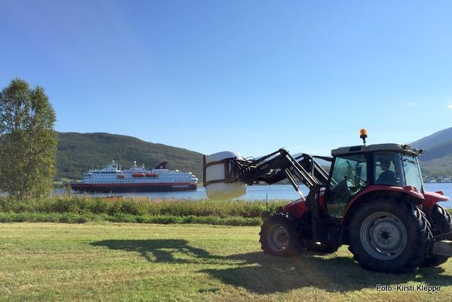 En traktor flytter høyballer på en åker mens Hurtigruta passer sundet i bakgrunnen - Klikk for stort bilde